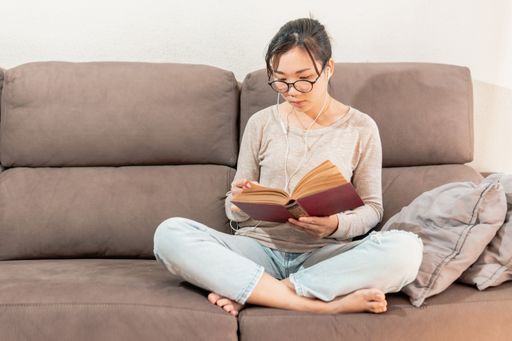 A young Asian woman sitting on the couch and reading a book while listening to music