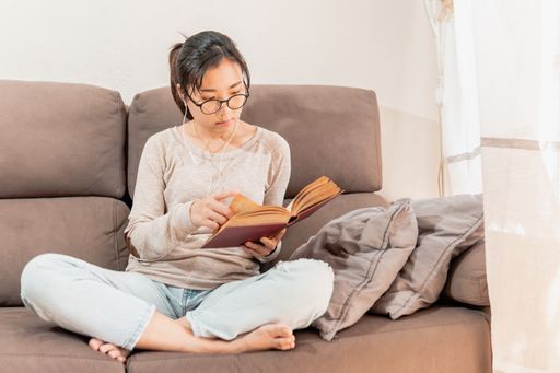 A young Asian woman sitting on the couch and reading a book while listening to music