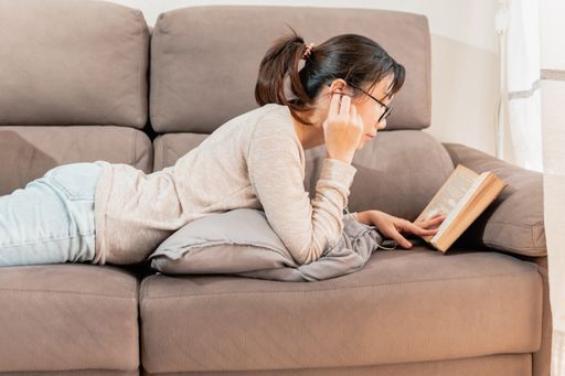 A young Asian woman lying on the couch and reading a book while listening to music at home
