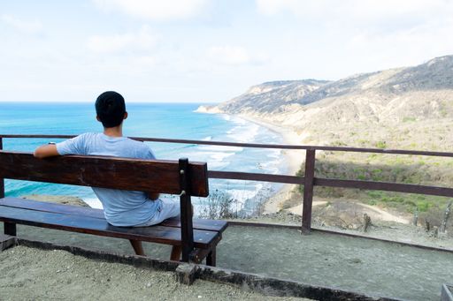 A young person sitting on the bench and looking at the sea surrounded by hills at daytime