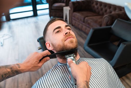 A closeup of a young hairdresser shaving the male customer's beard with a comb and a hair clipper in a barbershop