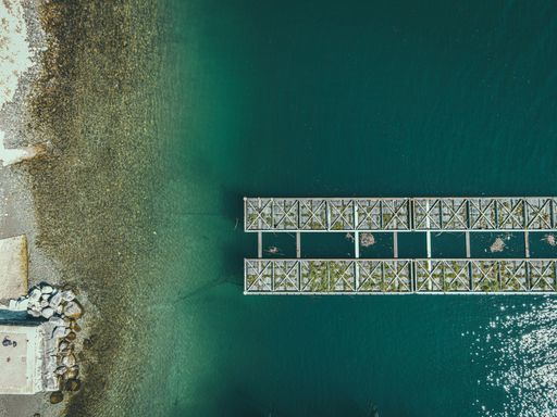 Aerial view of a turquoise shore with a metal pier over calm water, blending natural and industrial elements.