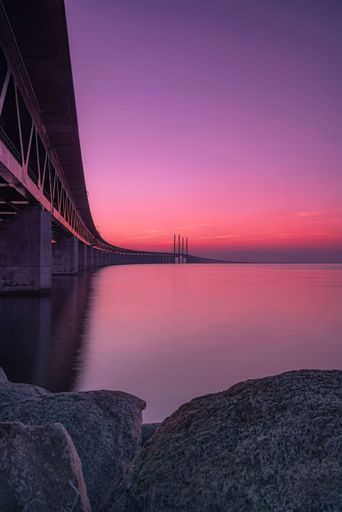 A vertical shot of a bridge above a lake captured from a rocky shore during a mesmerizing sunset