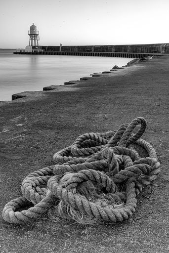 A vertical grayscale shot of a rope at the coast on the background of a lighthouse near Helsingborg, Sweden
