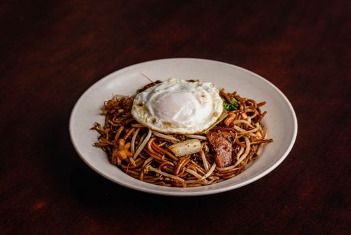 A closeup shot of a stir fried noodles with sunny side egg on top in a white plate on a wooden table
