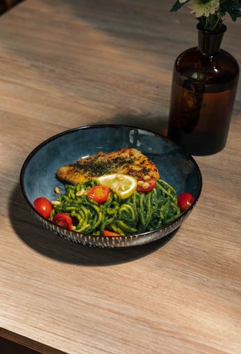 A closeup shot of a fried chicken breast and cooked vegetables in a dark turquoise bowl, with glass vase on a wooden table