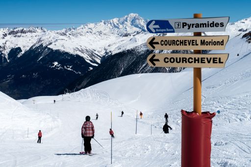 A closeup shot of direction signs on a skiing mountain filled with people and a mountain in the background
