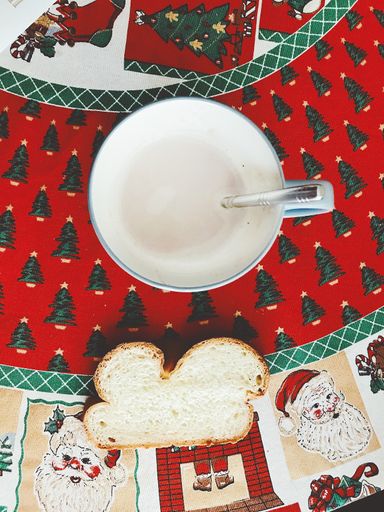 A vertical high angle shot of a cup of chocolate milk and a slice of bread on a Christmas-themed table clothing