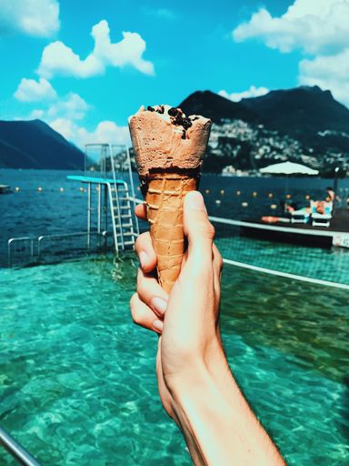 A vertical shot of a person holding a chocolate ice-cream cone with the beautiful sea and mountains in the background