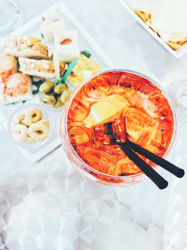A vertical high angle shot of a glass of alcoholic drink with ice cubes and straws near a tray with food