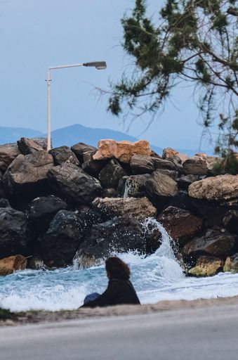 A vertical shot of a  woman sitting on the beach and watching the waves crash on nearby rocks