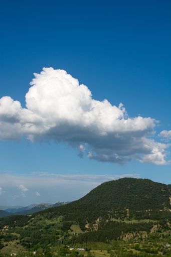 A vertical shot of a hill covered in greenery under a blue cloudy sky and sunlight