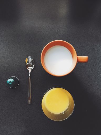 A vertical high angle shot of a cup of orange juice and milk beside a silver spoon on a black surface