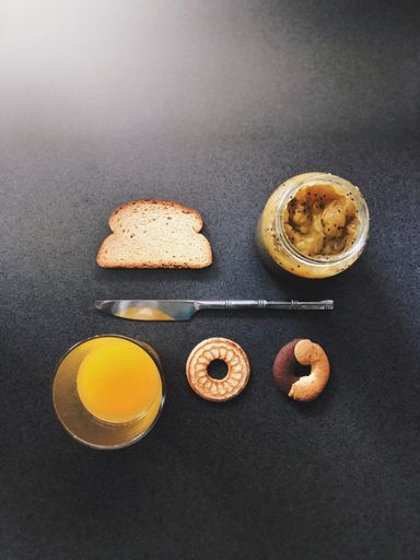 A vertical high angle shot of a knife between a slice of bread with peanut butter and an orange juice with cookies