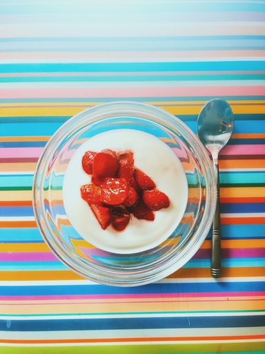 A vertical high angle shot of yogurt in a glass bowl with strawberries on top beside a silver spoon on a colorful striped surface