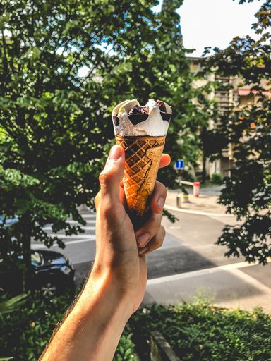 A vertical closeup shot of a person holding a chocolate and vanilla flavored ice-cream cone with green trees in the background