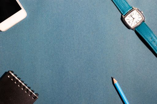 A pencil, smartphone, watch and a small notebook on the four corners of a  blue surface