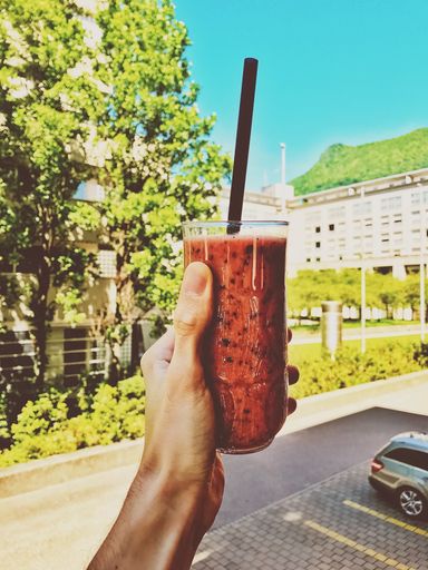 A vertical shot of a person holding a glass of smoothie with green trees and apartment buildings in the background
