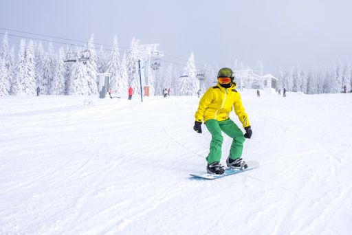 A snowboarder in a yellow and green costume riding down the slope with a ski lift in the background