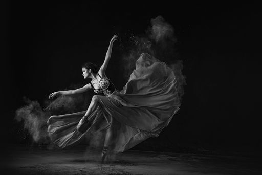 A grayscale shot of an emotional Southeast Asian ballet dancer in a dress with powder performing a move on a black background