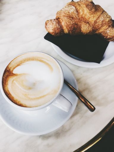 A vertical high angle shot of a cup of cappuccino and a croissant on a white surface