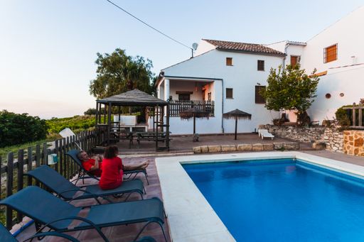An outdoor pool with people resting on chairs on a side in the back of a villa - rural houses for rent