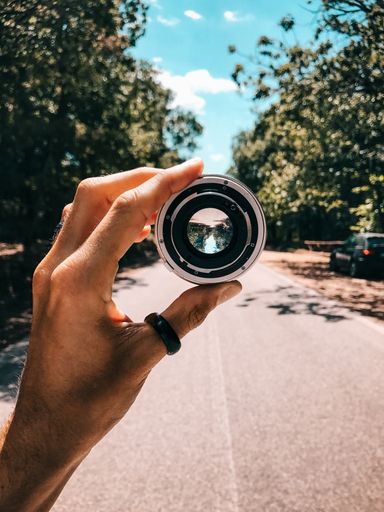 A vertical shot of a person holding a camera lens in the middle of the road surrounded by green trees