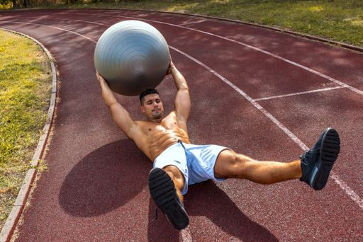 A fit shirtless man exercising with a gray ball on the floor in a park