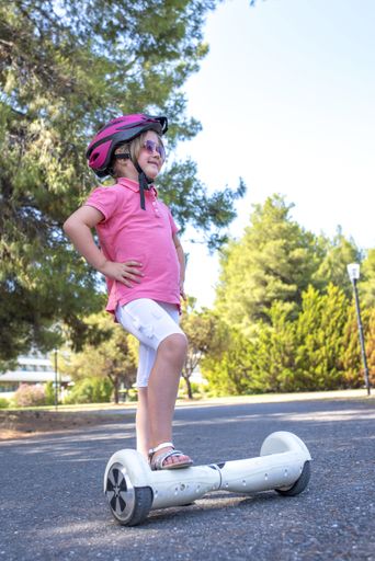 A vertical shot of a female child wearing a pink helmet standing posing with one foot on an electric skateboard in the street