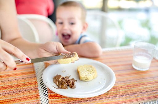 A closeup shot of female hands spreading a chocolate cream onto bread with an excited male child in the background