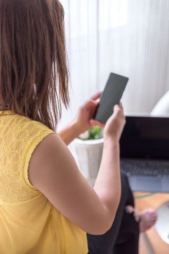 A vertical shot of a woman sitting on a chair holding her phone with a laptop  on a table near the window
