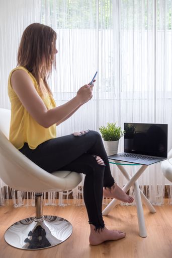 A vertical shot of a woman sitting on a chair holding her phone with a laptop  on a table near the window