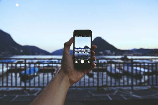 A person with a smartphone taking a picture of the breathtaking view with mountains and boats behind metal railings