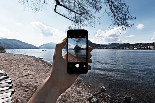 A person with a cellphone taking pictures of the beautiful lake near bare trees under the cloudy sky