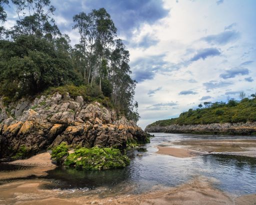 A landscape of Guadalmina Beach surrounded by rocks covered in greenery under blue cloudy sky in Asturias, Spain