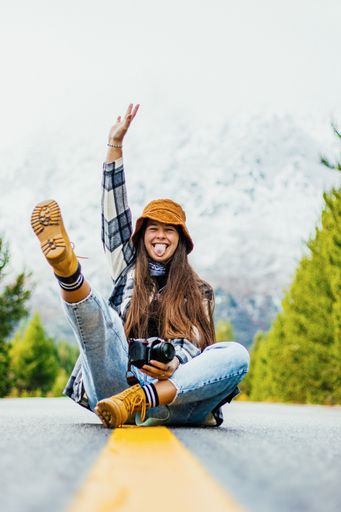 A vertical view of an attractive female photographer with a camera sitting on an asphalt road in a funny pose
