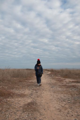 A vertical shot of a kid wearing warm clothes and walking alone on a field in cloudy sky background