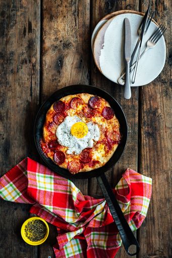 Homemade sourdough breakfast pizza baked in a cast iron skillet with pepperoni sausage and fried egg. On wooden surface shot from above