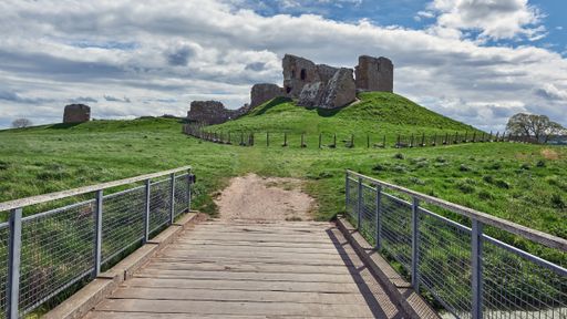Narrow bridge through green valley leads to the ruins of Duffus Castle, located on grassy hill near Moray, Scotland, under blue cloudy sky