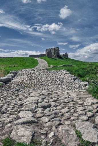 Narrow stone path through green valley leads to the ruins of Duffus Castle, located on grassy hill near Moray, Scotland, under blue cloudy sky