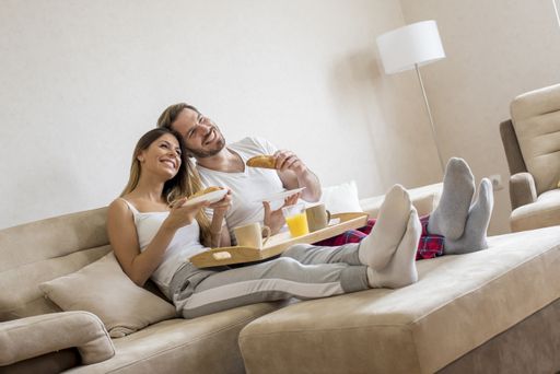 A cheerful couple eating breakfast on the sofa and watching TV under the lights