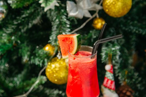 A horizontal closeup shot of a red cocktail in a glass with watermelon and Christmas tree in the background