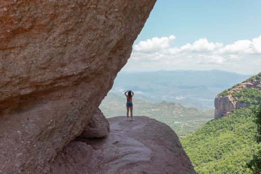 A back view of a young female wearing a red shirt and jeans shorts posing in front of scenic green hills on a sunny day