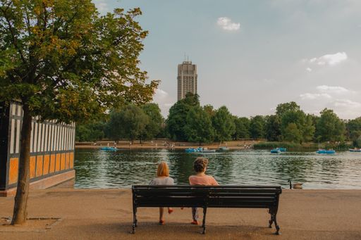 A view of male and female sitting on bench near lake in background of trees in Regent park