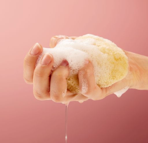 A female hand squeezing water out of a natural sea sponge body care on a feminine pink background