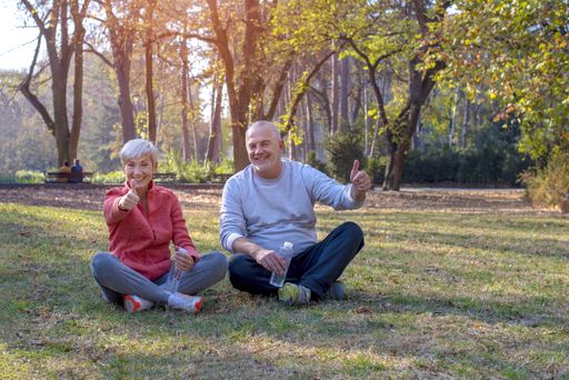 A senior couple sitting on the grass in a park happily, holding their thumbs up during daylight