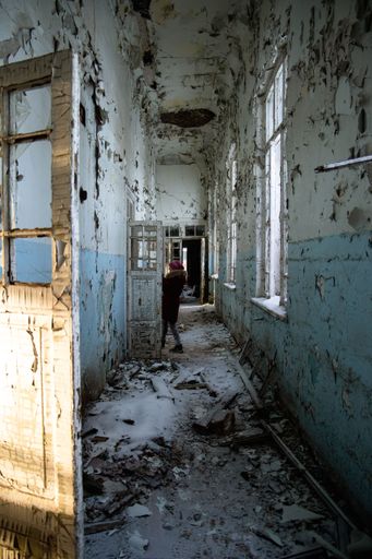 A woman standing in a corridor of an abandoned building with damaged walls, doors and windows