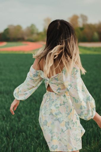 A vertical shot of a Caucasian female in a dress seen from behind, walking on a green field