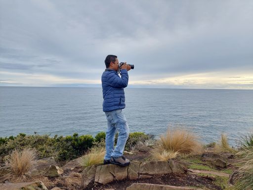 A male hiker in a blue jacket taking professional photos of a cliff over a seashore