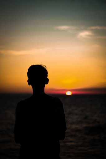 A vertical back view of a person's silhouette watching the sun setting in the beautiful golden sky